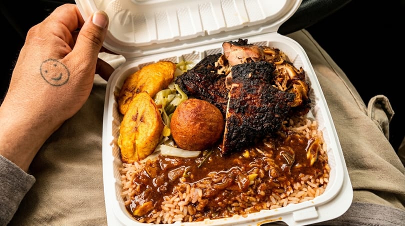 A hand with a smiley face tattoo holds a takeout container of jerk chicken, rice, plantains, and a dumpling.
