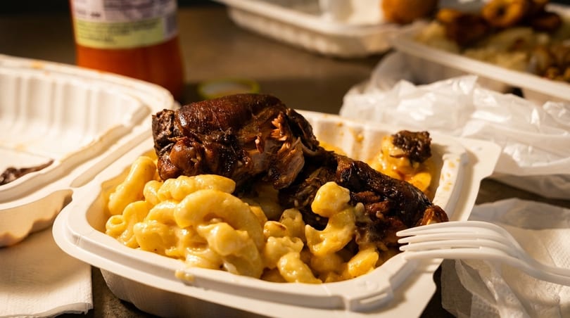 Takeout container with mac and cheese and a large piece of roasted meat, with a fork.